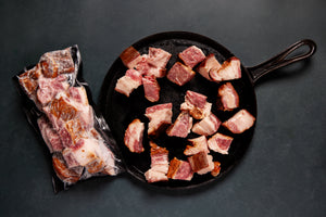A package of Wagyu Beef Bacon Ends next to a skillet with beef bacon ends scattered inside. 