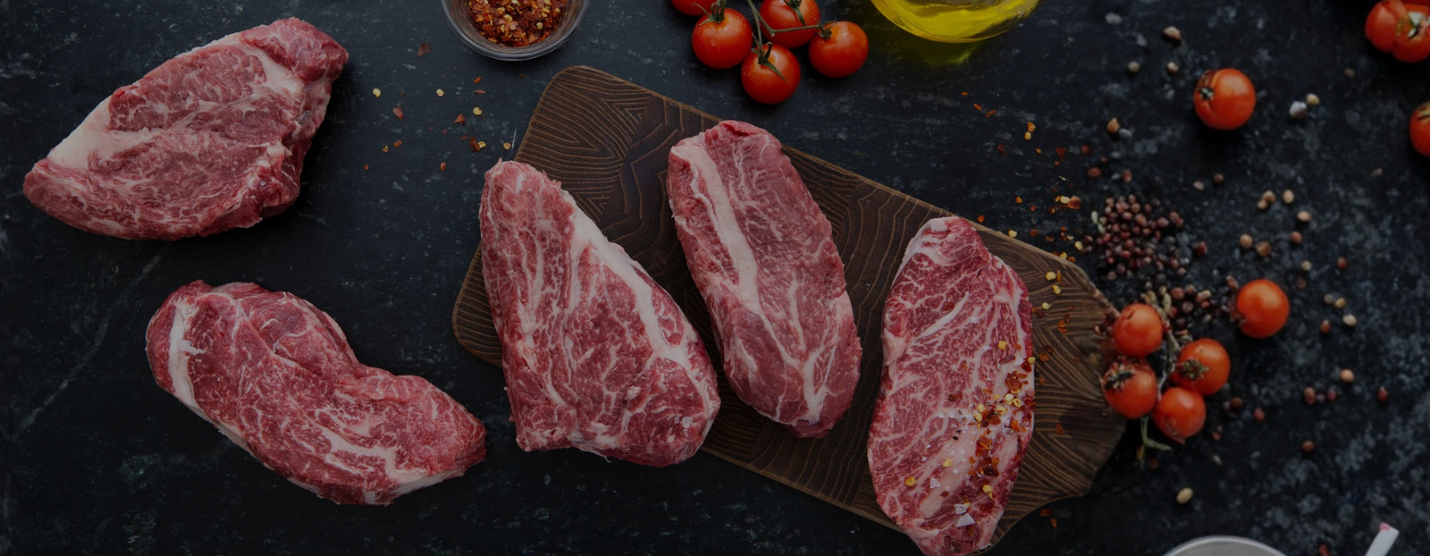 five deeply marbled wagyu chuck eye steaks laying across a cutting board and slate background with tomatoes and peppercorns 