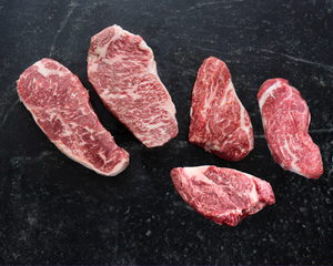 Two raw Wagyu NY Strips and three raw Wagyu chuck eyes on a black slate counter