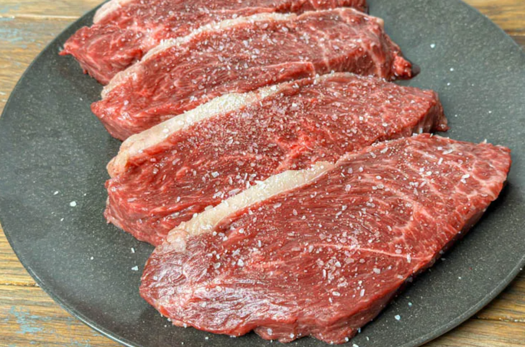Four Raw Picanha Steaks on a grey plate with a dusting of salt.