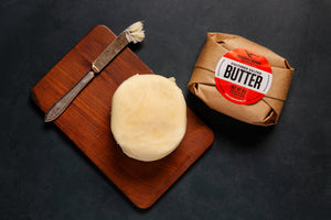 One wrapped Ploughgate butter next to a cutting board with an unwrapped butter and a butter knife with a pad of butter on the end