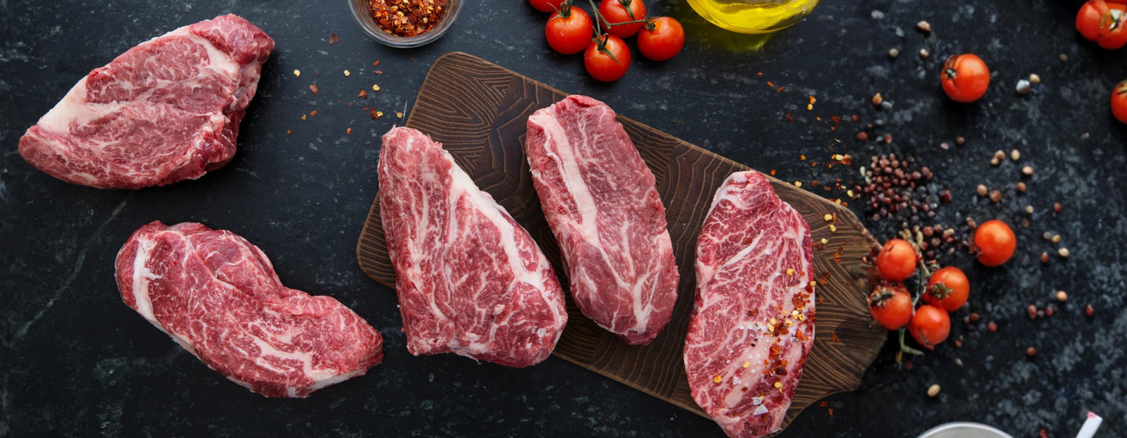 Five deeply marbled Wagyu Chuck Eye steaks on a cutting board surrounded by tomatoes on the vine, pepper corns, and red pepper flakes. 