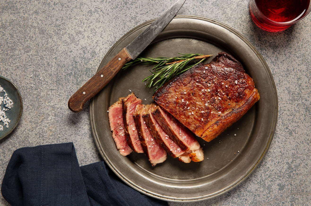 A cooked Vermont Wagyu 100% Fullblood Wagyu NY Strip Steak half sliced on a silver plate with a steak knife next to it. 