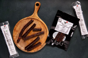 A package of Vermont Wagyu snack sticks, a single package of Jalapeno Cheddar stick, a single package of maple whiskey stick, and a cutting board with snack sticks. 